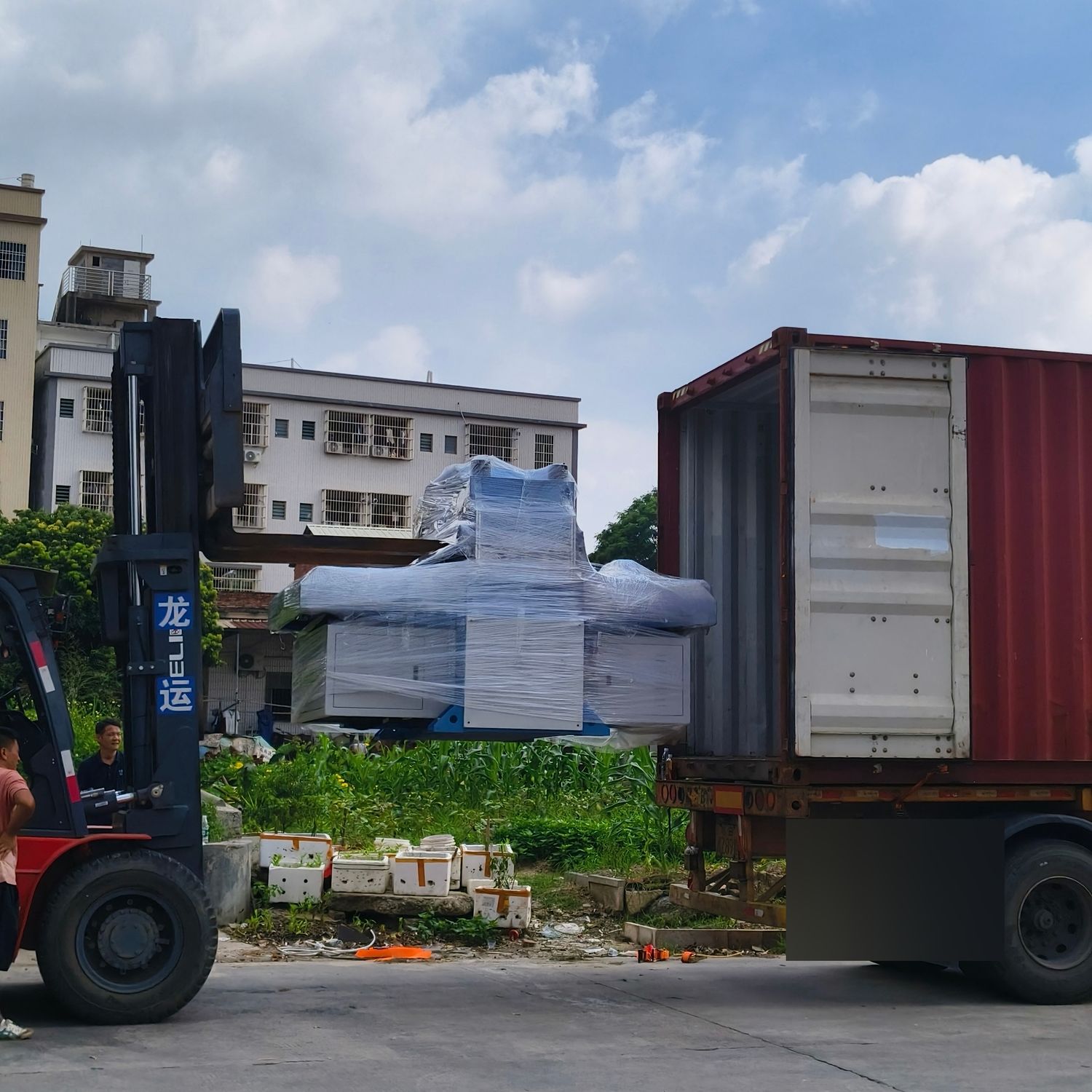 Forklift feeding a wrapped plastic machinery unit into an open container