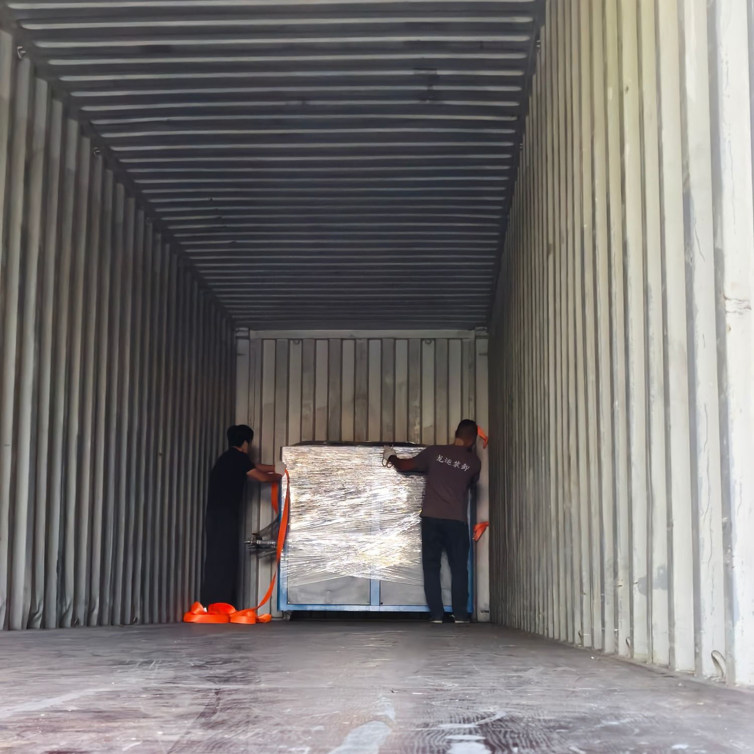 Workers lashing a wrapped machine with ratchet straps inside a shipping container